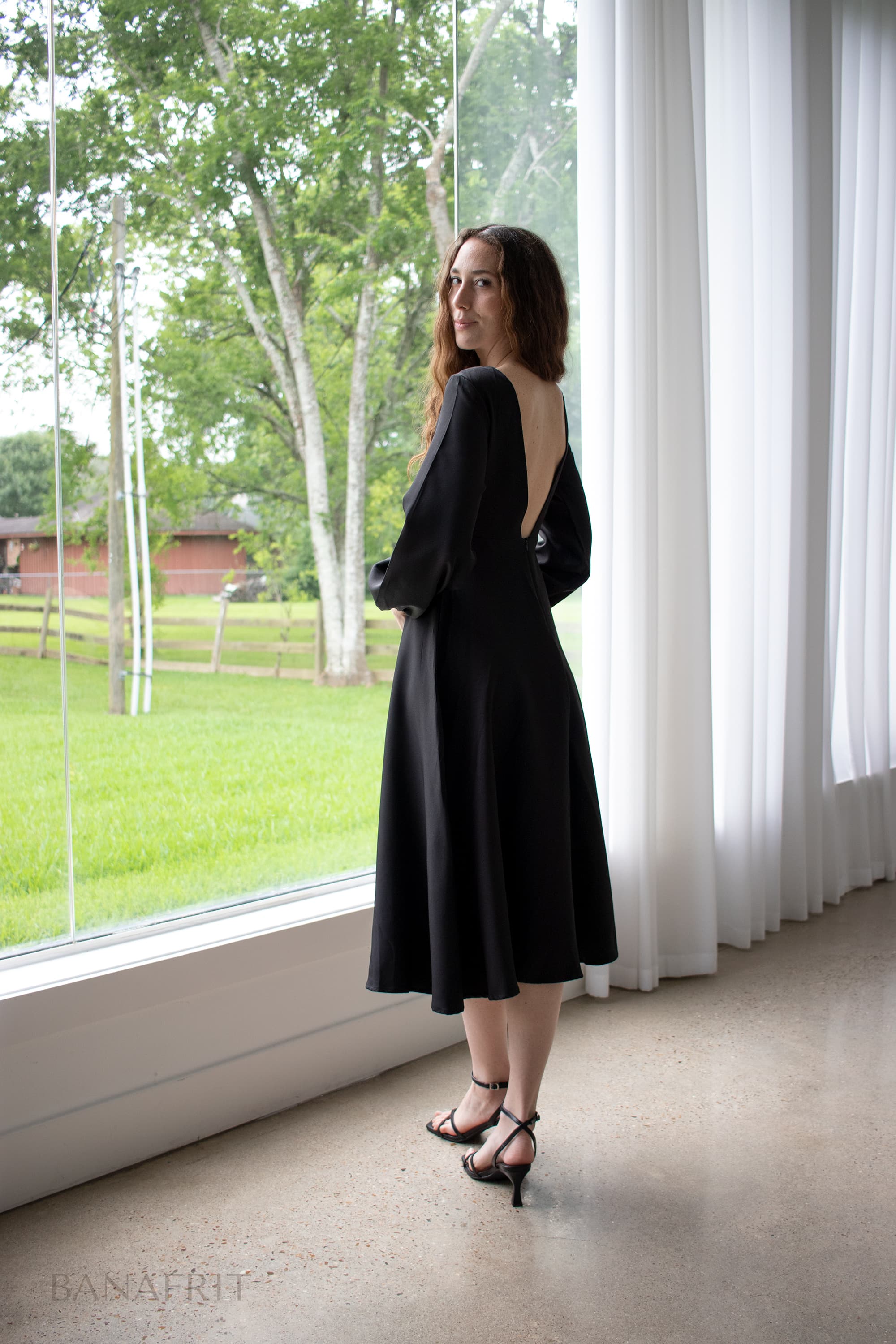 Fine art portrait — woman in black off-shoulder dress, standing before panoramic window, natural light with her back turned.