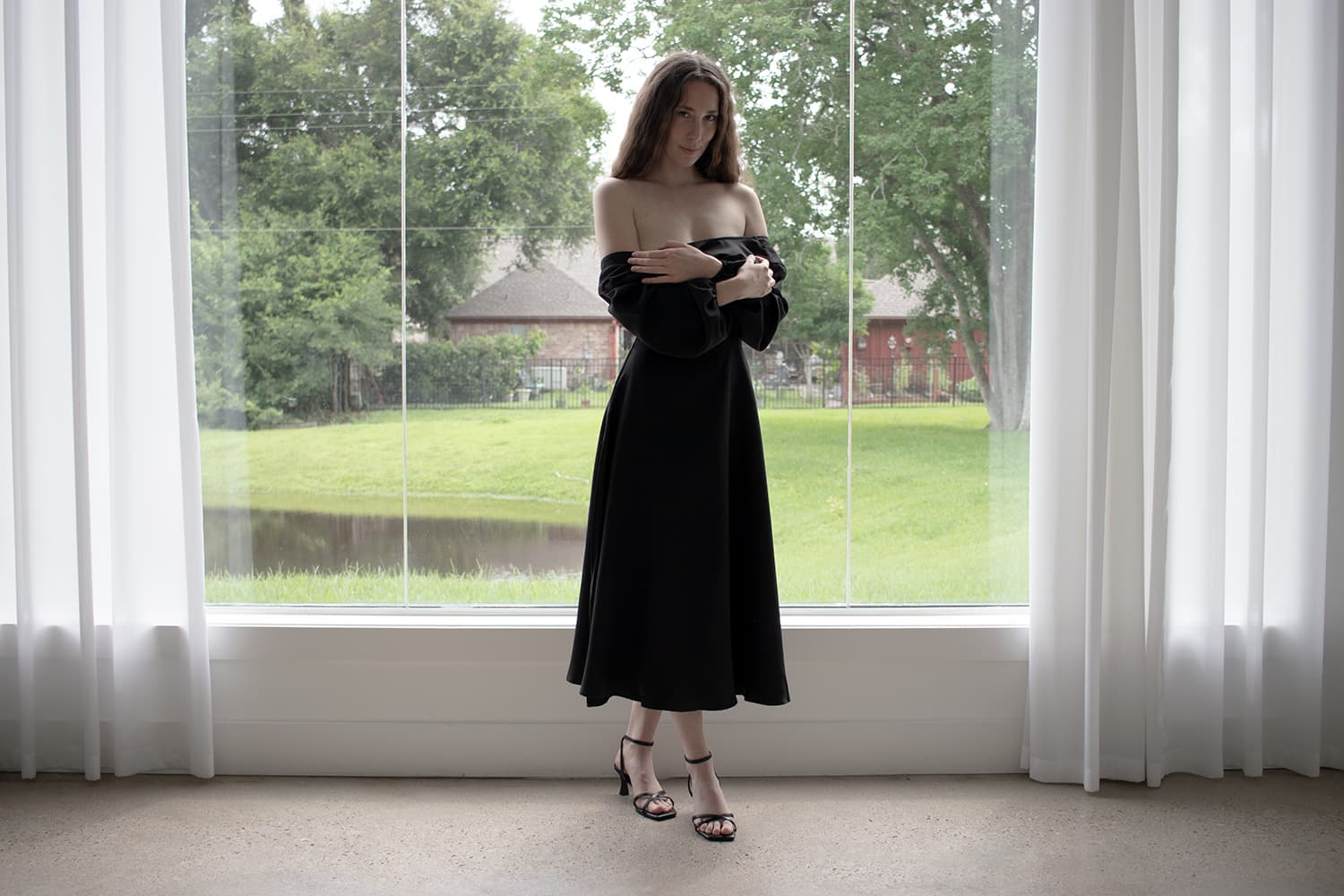 Fine art portrait — woman in black off-shoulder dress, arms crossed, standing before panoramic window, natural light