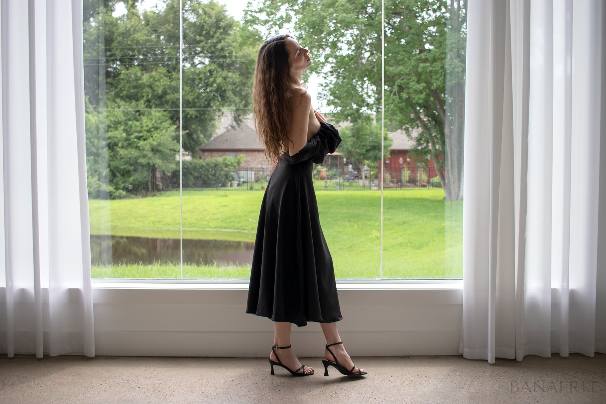 Fine art figure study — woman in black dress, back to camera, gazing upward before large window, soft overcast light