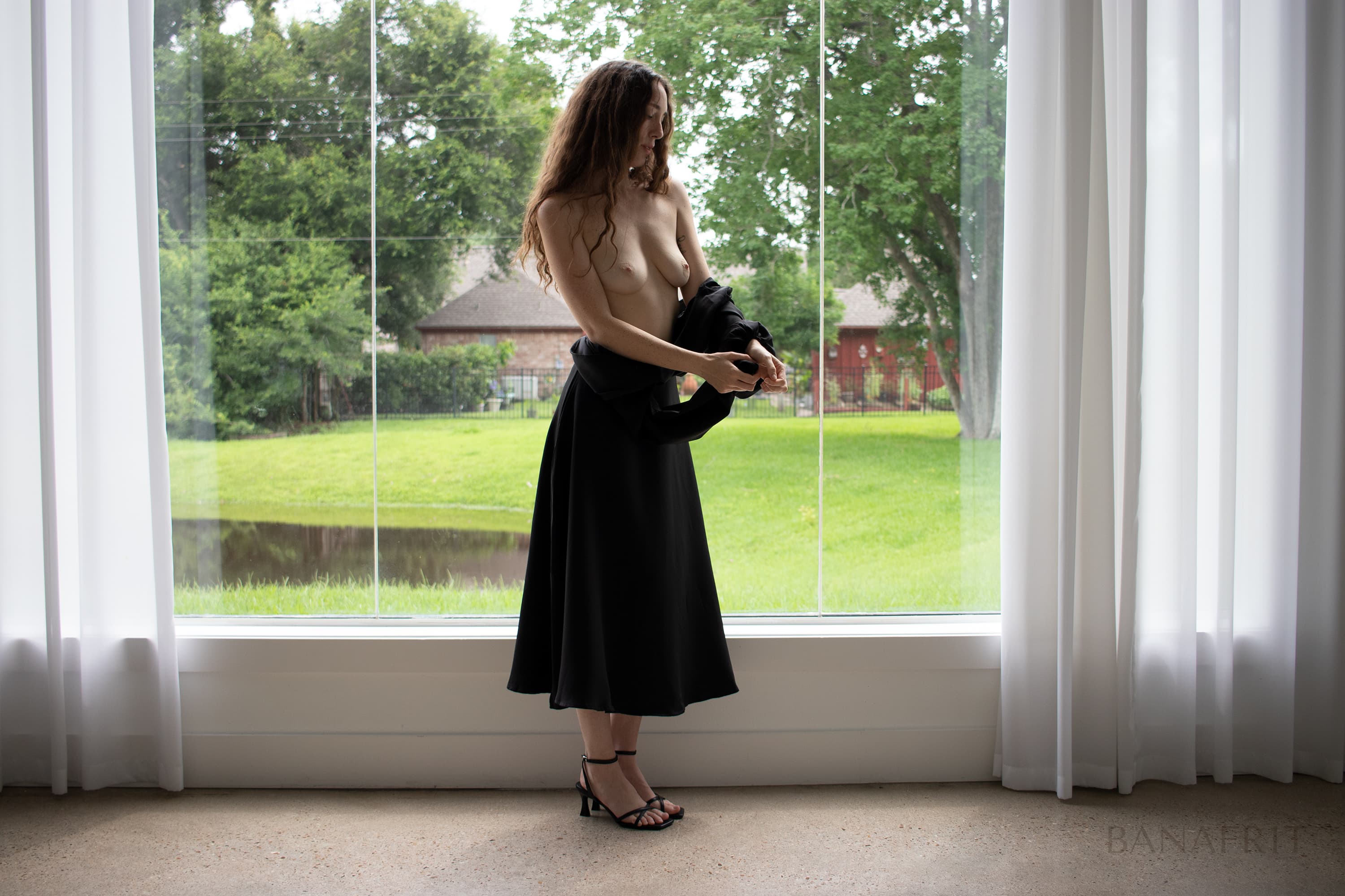 Fine art figure study — topless woman removing her black dress, before large window, soft overcast light