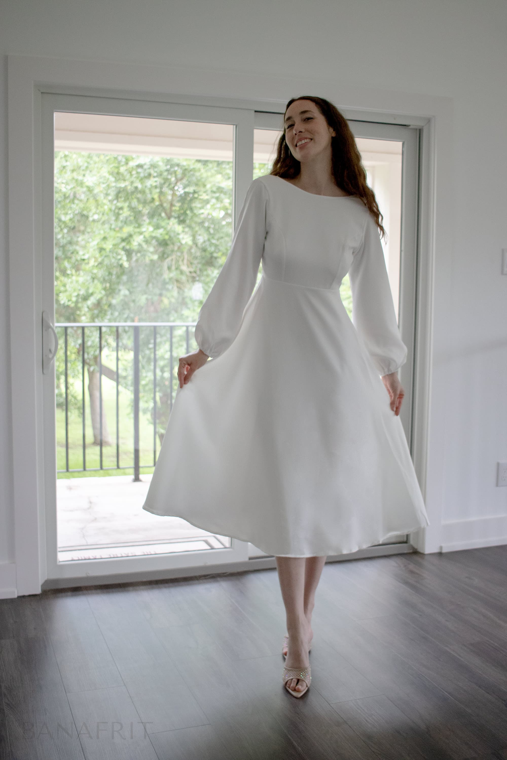 Fine art portrait — woman in white long-sleeve dress, mid-movement, natural light through glass door